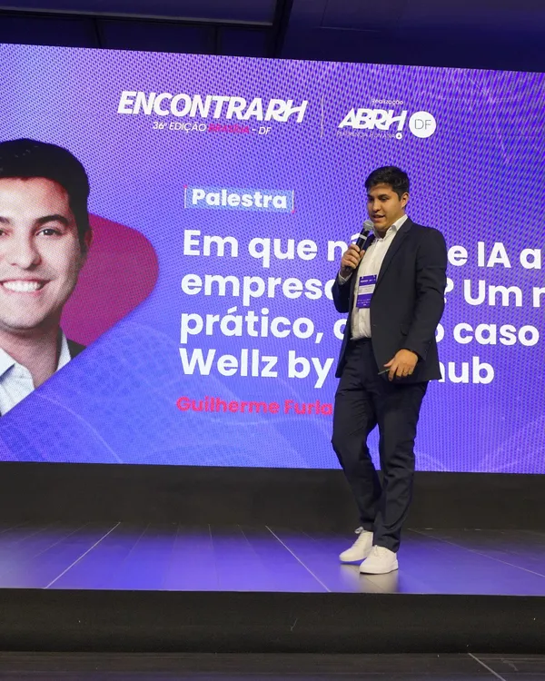 Guilherme Furlan de terno, com microfone, no palco do 36º EncontraRH, diante do telão com o título da palestra